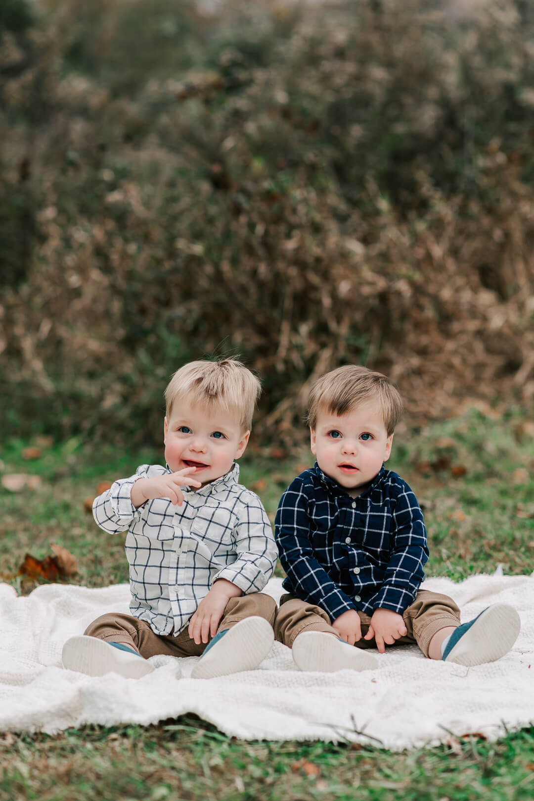 Two young toddler boys sit on a picnic blanket in a park