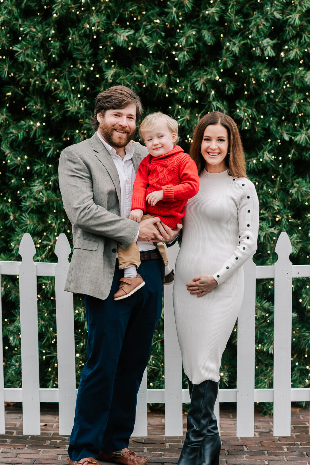 A pregnant mom stands against a white picket fence with her husband holding their toddler son