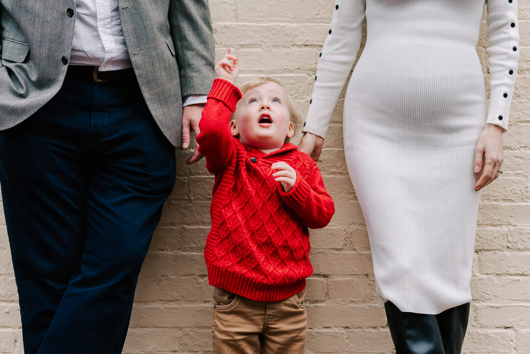 A toddler boy in a red sweater points up while standing against a white brick wall between mom and dad after they used fertility clinics fairfax va
