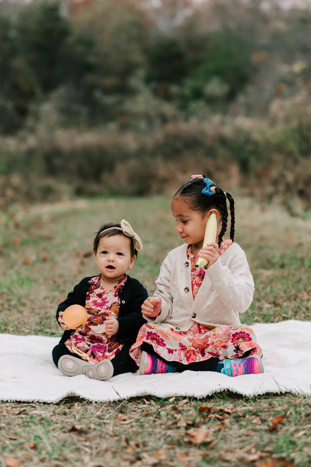 Two toddler sisters in matching pink floral dresses sit on a picnic blanket in a park playing with fruit