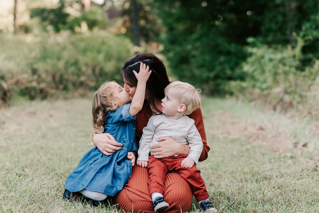 A mother kisses her toddler daughter while her toddler son sits on her lap