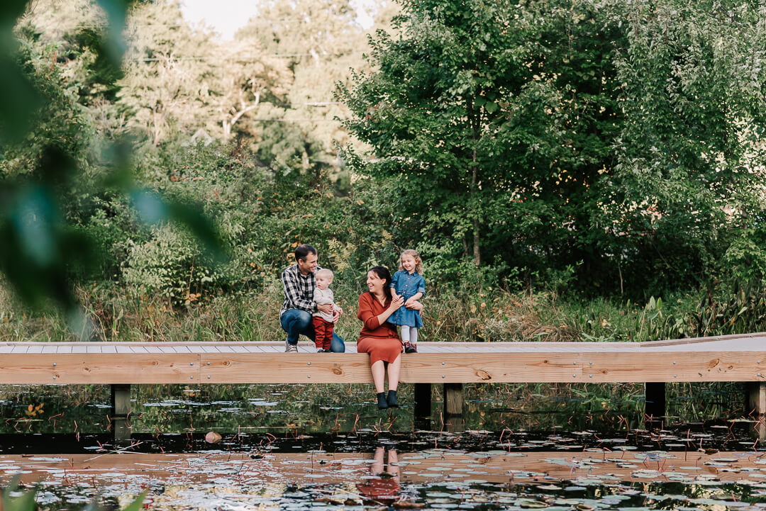 A mom and dad sit on a boardwalk over a pond with their two toddler children after visiting a mclean preschool
