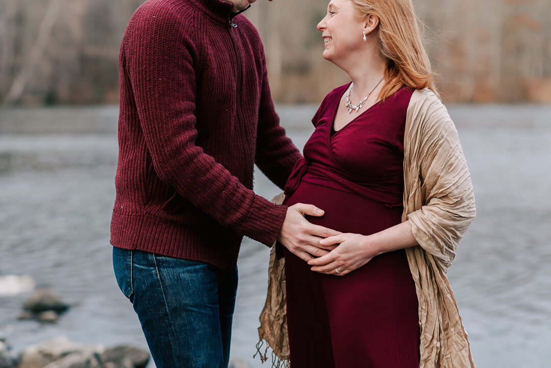 A mother and father to be hold the bump while standing in maroon by a lake