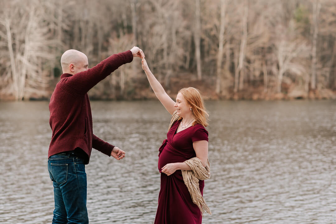 A father to be twirls his pregnant wife while dancing by a lake in maroon dress and sweater after meeting metropolitan doulas