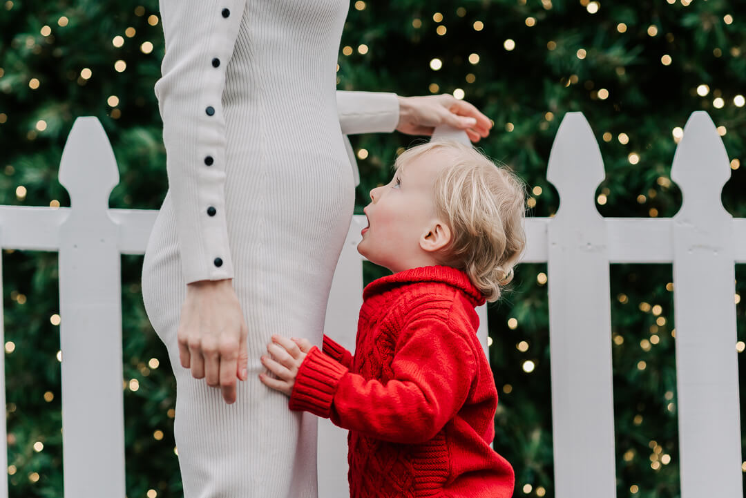 A toddler boy in a red sweater leans in to kiss mom's pregnant belly while standing in front of a white picket fence