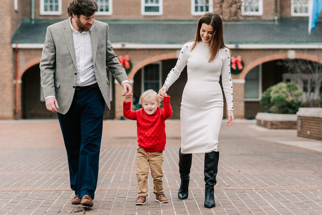 An expecting mother and father walk their toddler son in a red sweater through a brick patio holding hands after visiting birthing center arlington