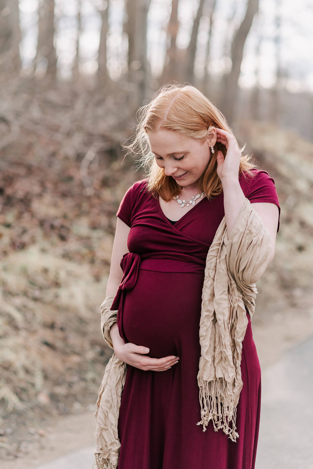 A mother to be smiles down to her bump while standing in a park path in a red maternity gown