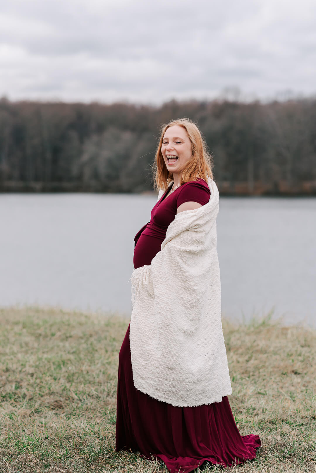 A happy mother to be laughs while walking along a lake in a red maternity gown and wrapped in a blanket after meeting doula arlington va