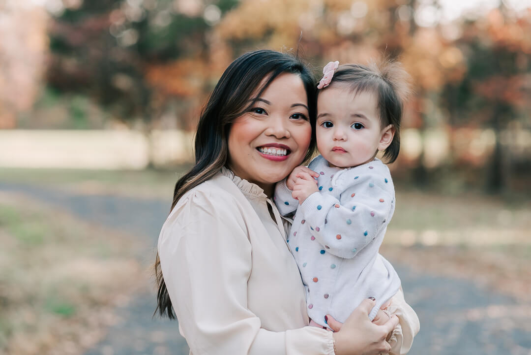 A mother smiles while standing in a park in fall with her toddler daughter on her hip