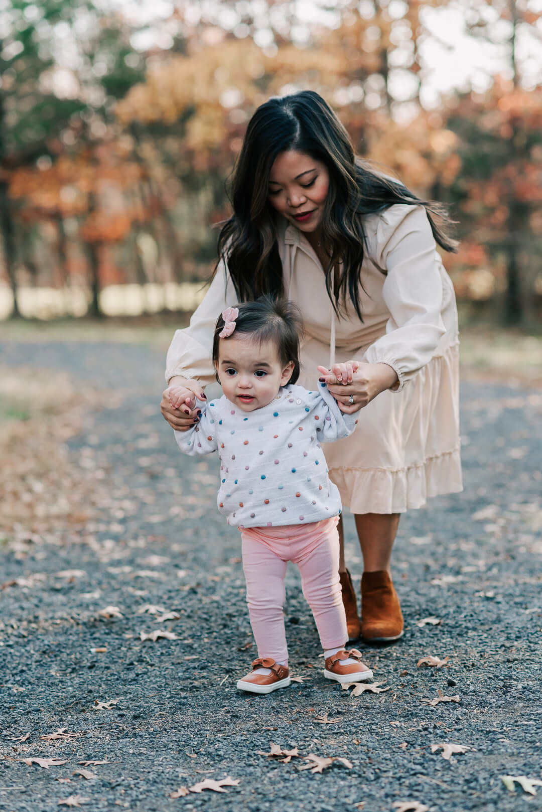 A mother holds the hands of her toddler daughter while they walk through a park path in fall after visiting birthday party places northern va