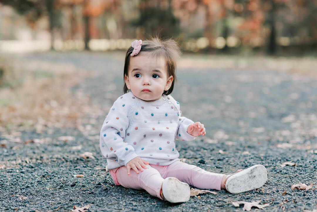 A toddler girl in pink pants and colorful sweater sits in a gravel path in a park before visiting birthday party places northern va