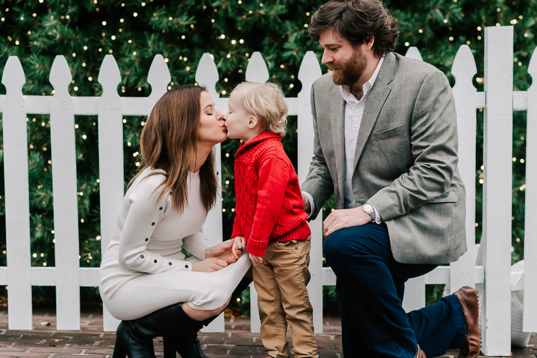 A mother in a white dress kisses her toddler son in front of a white picket fence with dad looking on after using birthing center washington dc