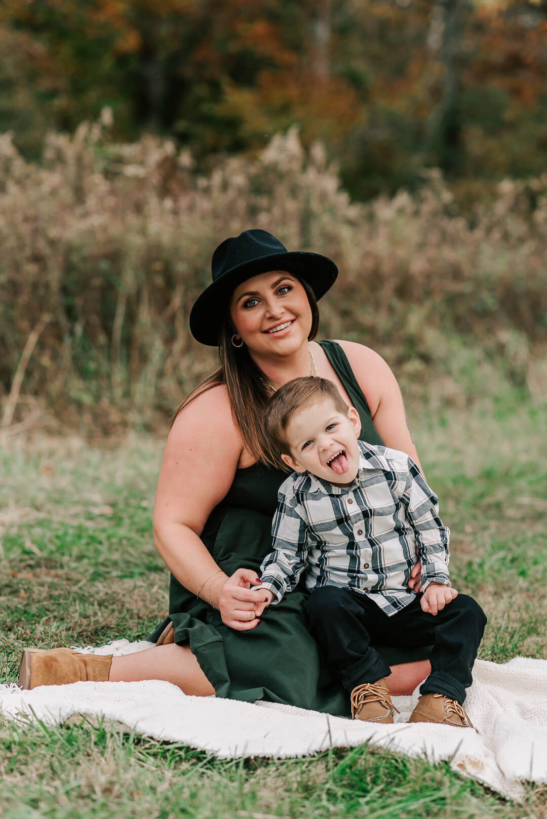 A mother in a black hat and green shirt sits in a park lawn with her toddler son making silly faces on her lap