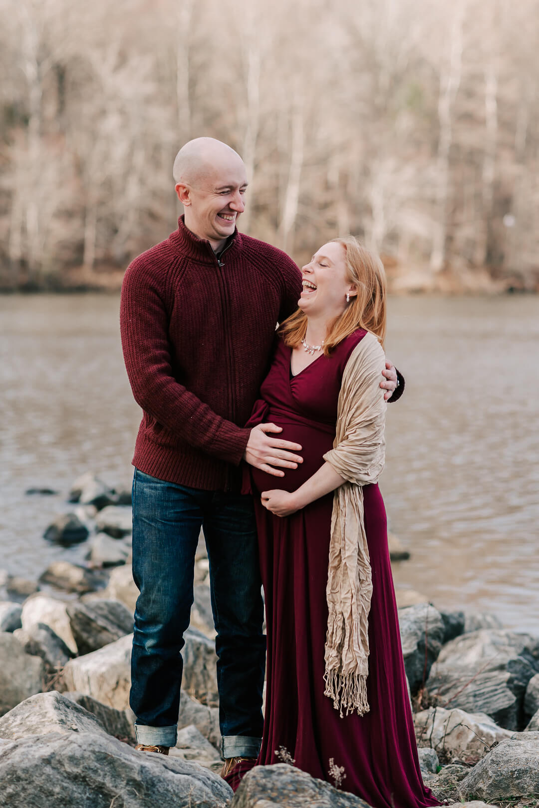 Happy expecting parents laugh while standing on a rocky riverside in maroon sweater and maternity dress