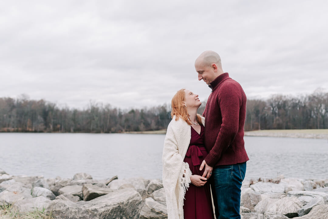 Happy expecting parents stand on the edge of a river holding the bump and smiling at each other after their 3d ultrasound northern virginia