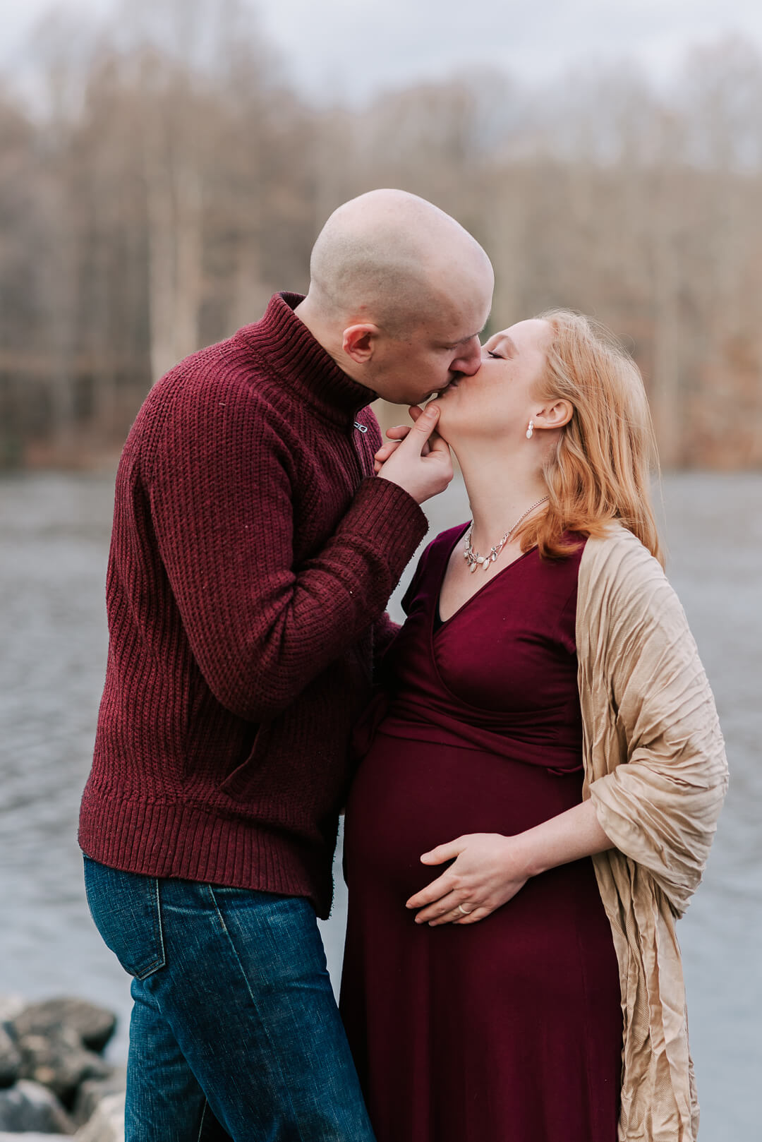 A mom to be kisses her husband in a maroon dress while standing along a river before their 3d ultrasound northern virginia