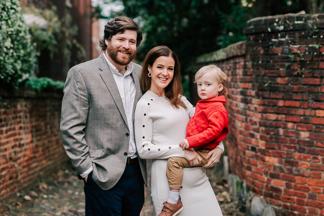 Happy parents stand in a brick wall alley with their toddler sitting on mom's pregnant bump