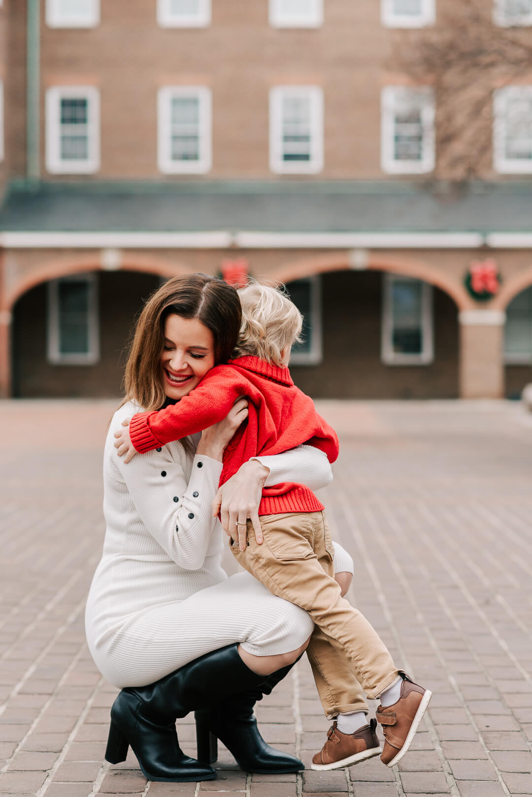 A happy mother in a white dress hugs her toddler in a red sweater on a brick patio thanks to fertility acupuncture northern virginia