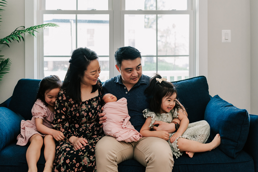 A mom and dad sit on a blue couch under a window with their newborn baby sleeping in a swaddle in dad's lap with their toddler daughters on either side before some mommy and me classes northern virginia