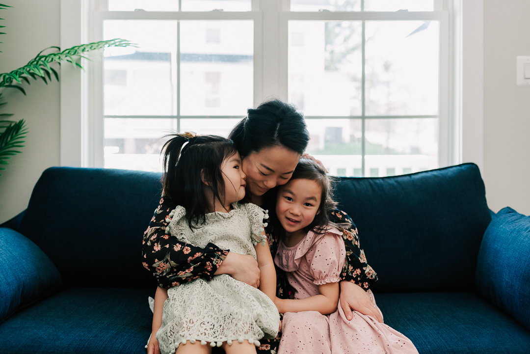 A happy mother sits on a blue couch under a window hugging her toddler daughters in her lap after some mommy and me classes northern virginia