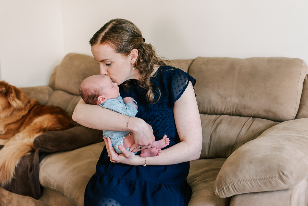A new mother kisses the head of her newborn baby in her arms while sitting on a couch with their dog thanks to northern virginia fertility clinics
