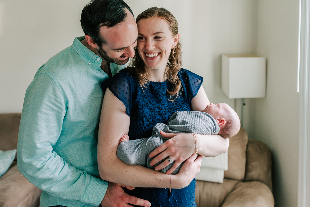Happy new parents snuggle while standing in a window and smiling with their sleeping newborn baby in mom's arms thanks to northern virginia fertility clinics