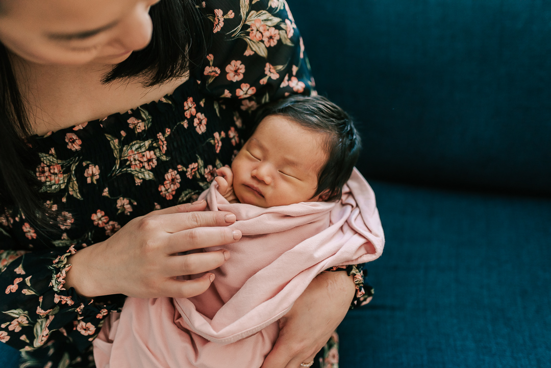 A mother in a black floral print dress sits on a blue couch cradling her sleeping newborn baby in a pink swaddle after meeting northern virginia lactation consultants