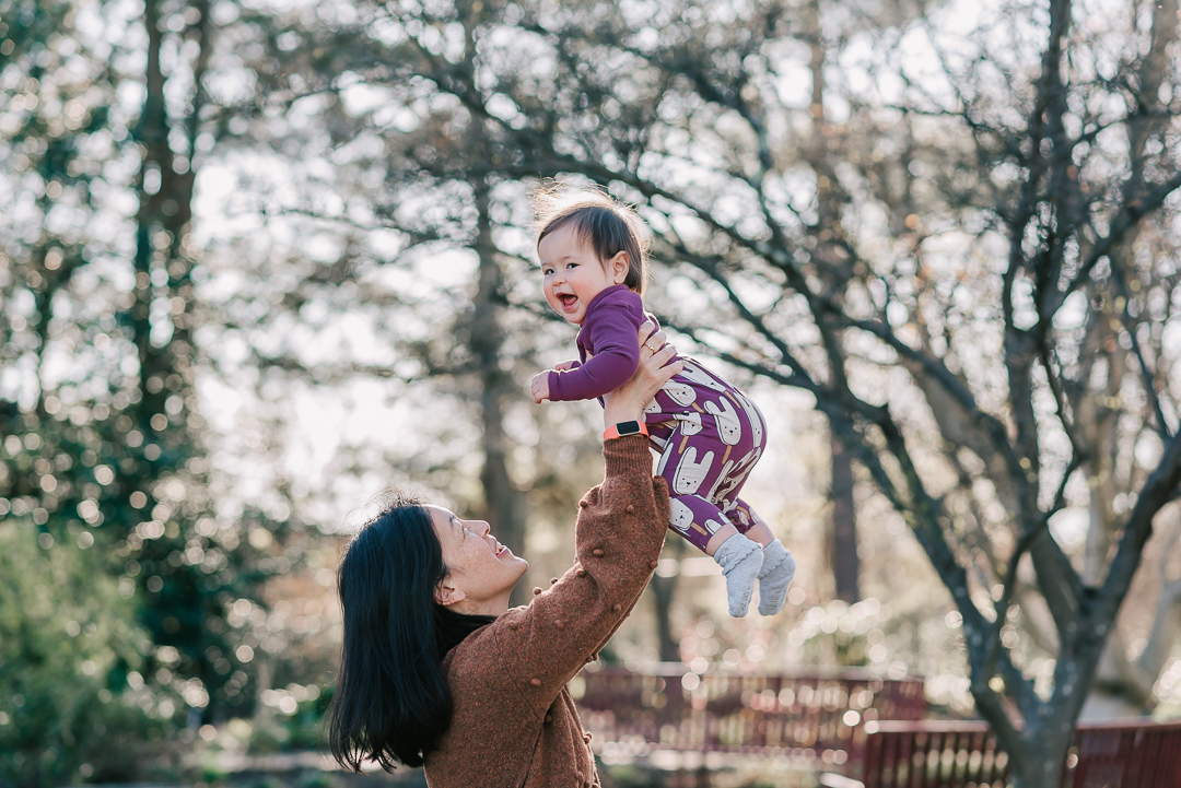 A happy mother in a brown sweater lifts her laughing toddler daughter above her head in a park