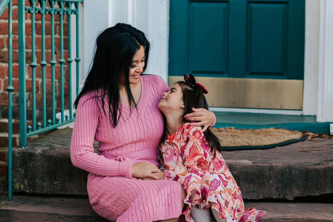 A happy mother in a pink dress sits on a porch step hugging her toddler daughter in a pink floral print dress