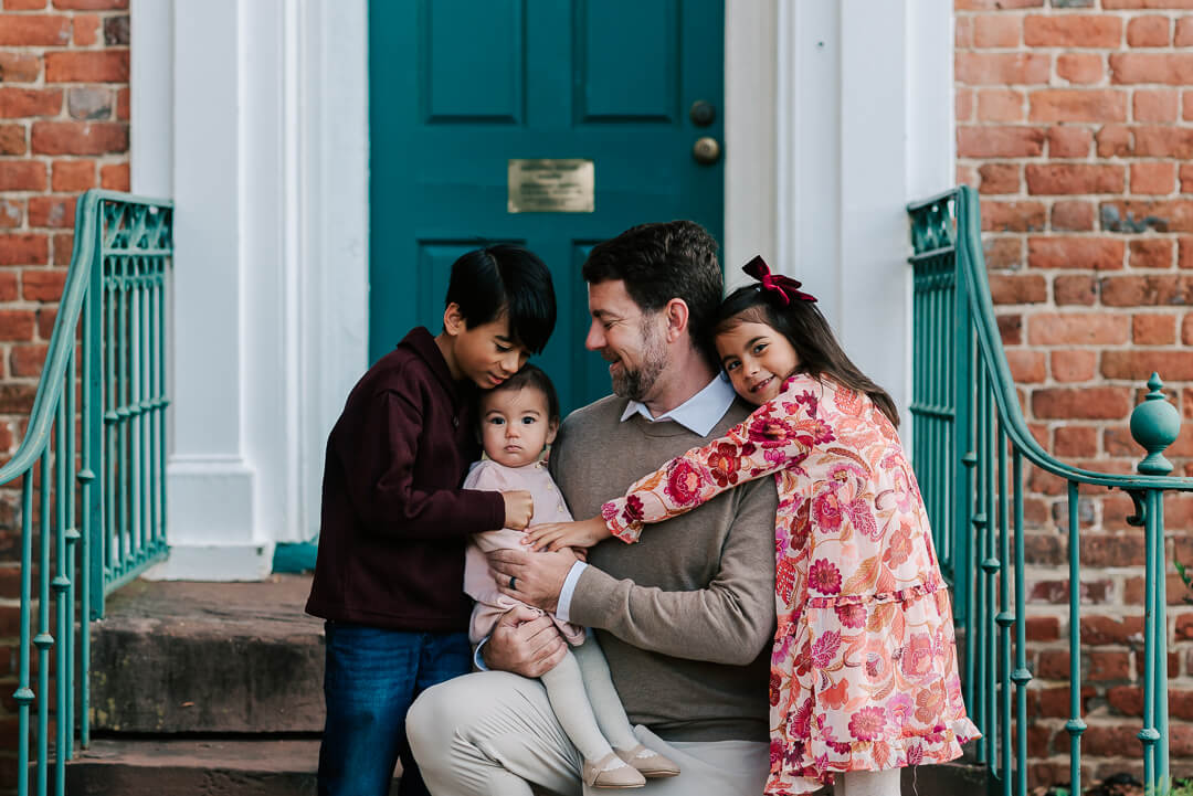 A happy father is hugged by his toddler daughter while holding his infant daughter in his lap with his son hugging her on a porch step before visiting northern virginia preschools