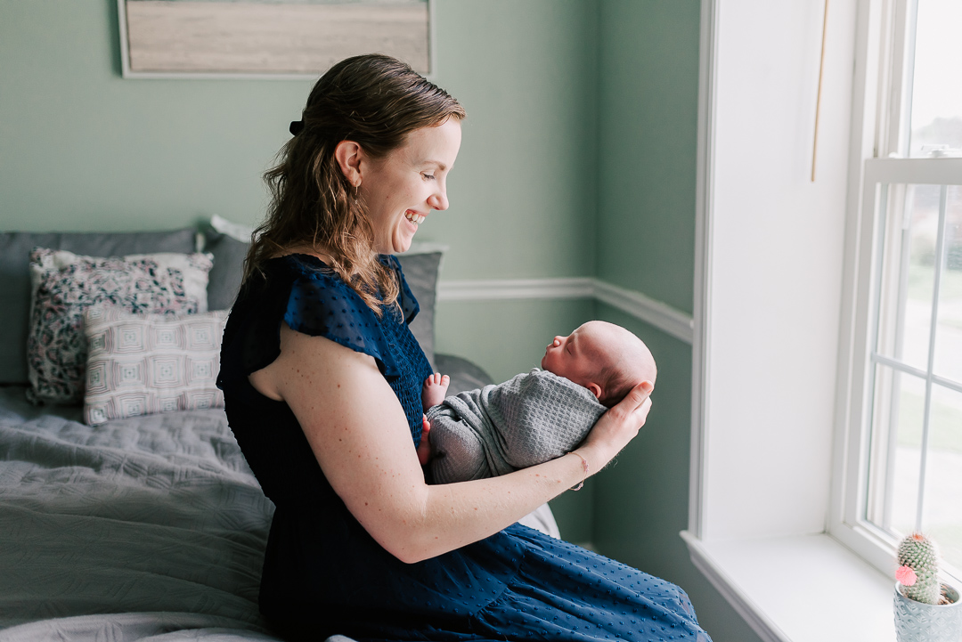 A happy new mother smiles down at her sleeping newborn baby in her hands while sitting on a bed in a window before some pelvic floor therapy northern virginia