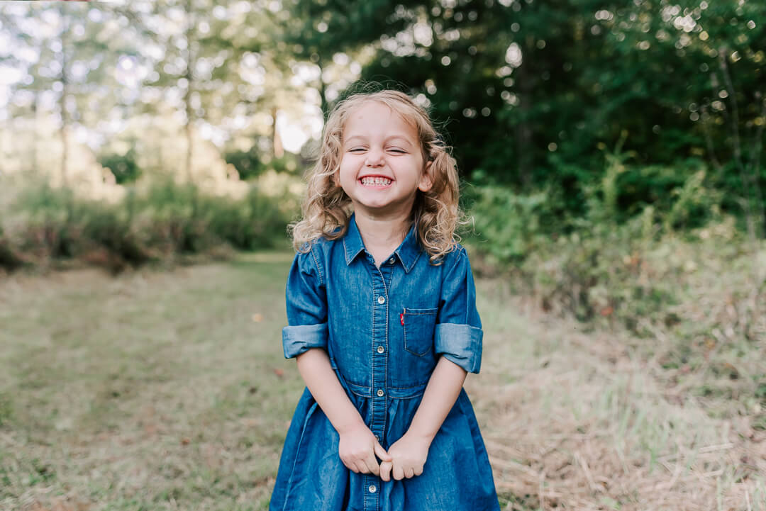 A happy toddler girl in a denim dress smiles big while standing in a park trail before visiting childrens museum northern virginia