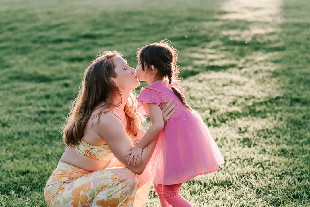 A mother and her toddler daughter share a kiss in a grassy lawn at sunset