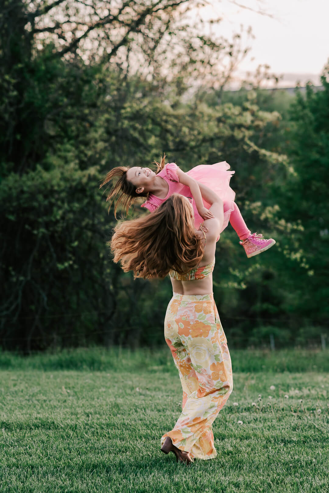A mom in colorful pants lifts and spins with her toddler daughter in a pink dress in a field after some family therapy northern virginia