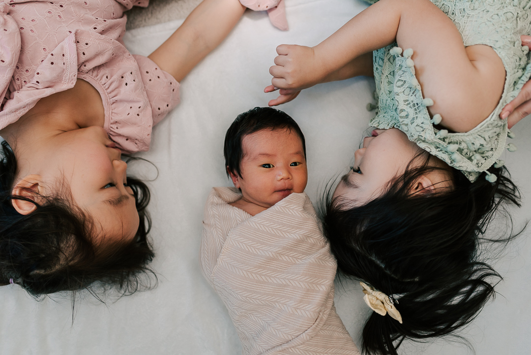 Toddler sisters lay on a bed with their newborn baby sibling wrapped in a brown swaddle