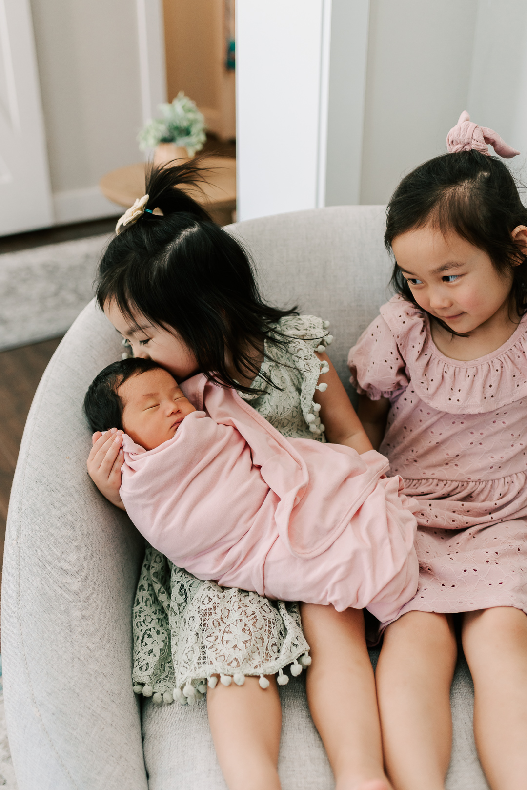 Toddler girls in pink and green dresses sit in a chair while one holds and kisses their newborn sibling before visiting private schools in northern virginia
