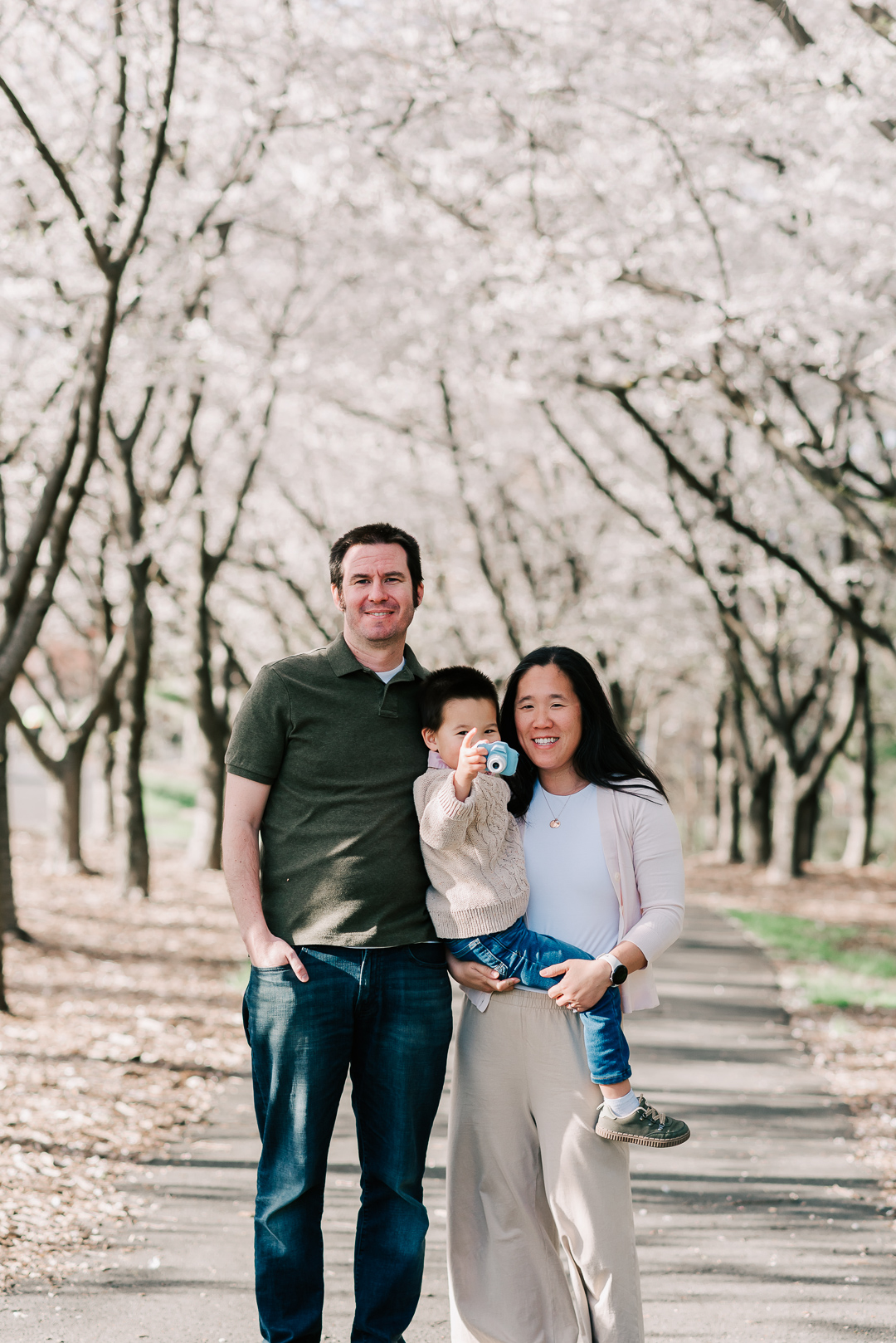 Happy mom and dad stand in a park sidewalk lined with flowering trees while carrying their toddler son with a toy camera before visiting swim lessons northern virginia