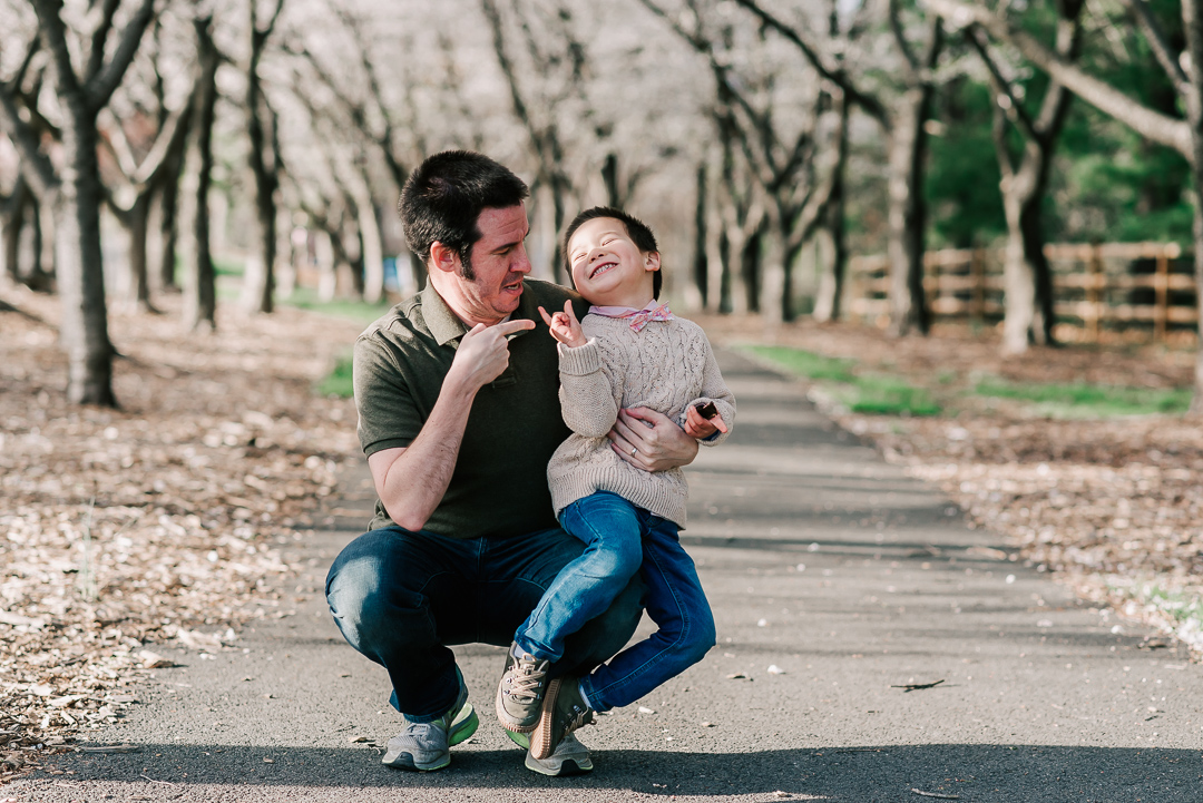 A father makes silly faces and points at his toddler son in his lap while in a park sidewalk at sunset before visiting swim lessons northern virginia