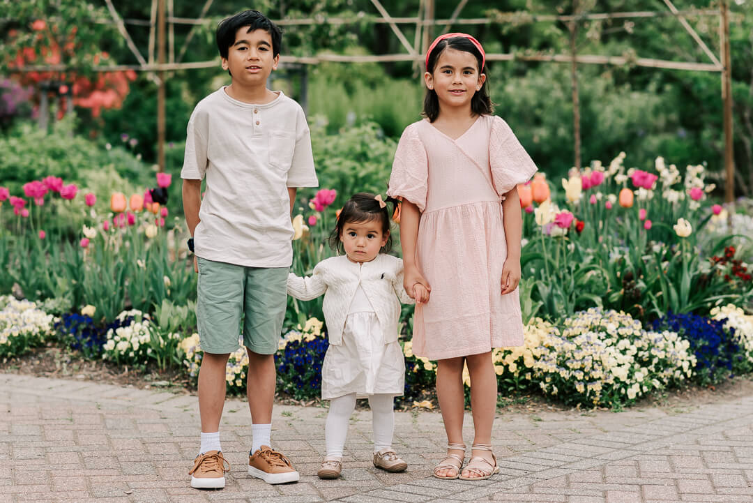 Toddler brother and sister stand in a garden path holding the hands of their younger sister in a white dress