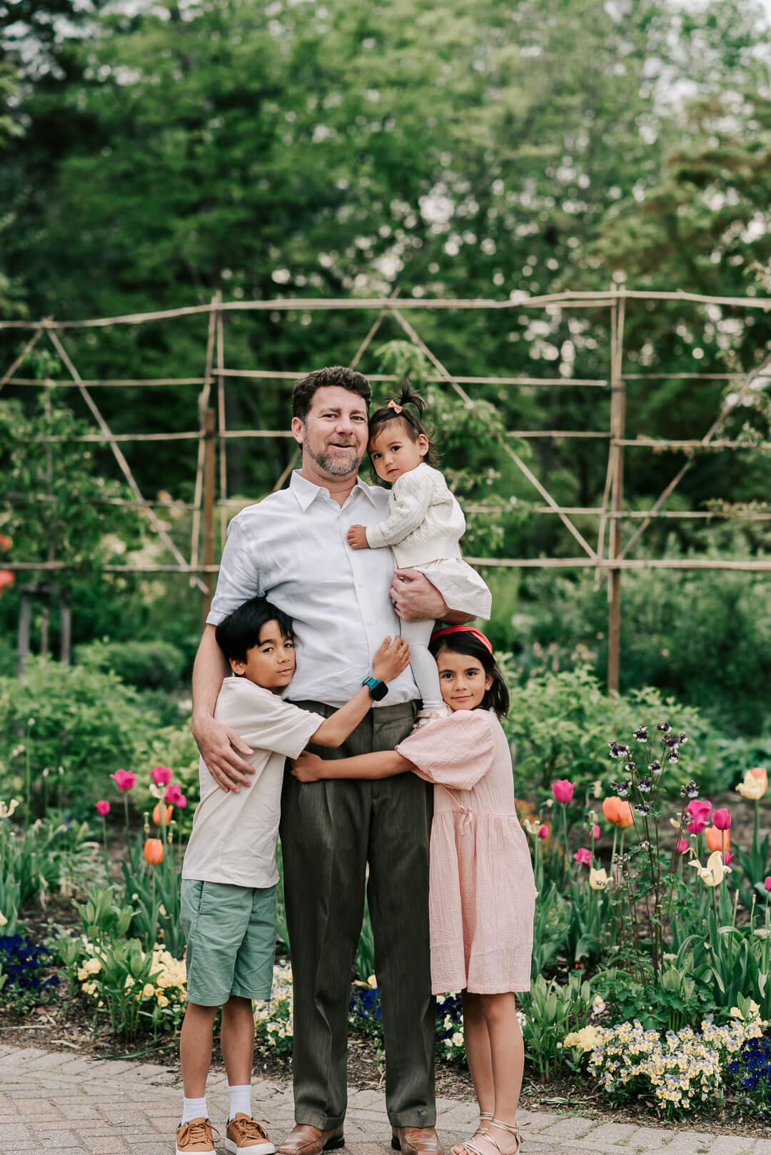 A happy father stands in a garden holding his youngest daughter while his toddler son and daughter hug onto him before visiting kid friendly restaurants northern va