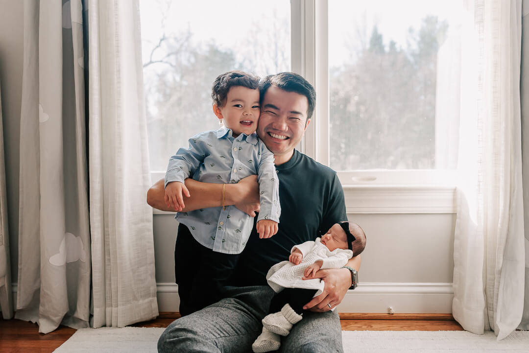 A smiling father hugs his toddler son against his cheek while holding his sleeping newborn daughter in his lap