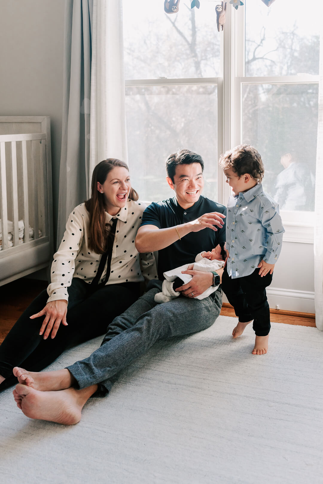 Happy parents laugh with their toddler son on the floor of a nursery while dad cradles their sleeping newborn baby before some kid friendly things to do in dc