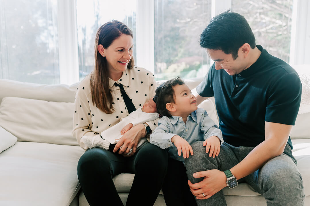 A toddler boy sits between mom and dad on a couch under a large window while mom holds a newborn baby before doing some kid friendly things to do in dc