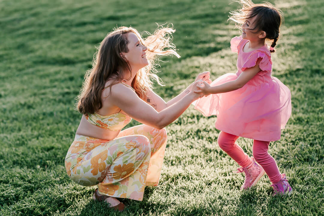 A laughing and smiling mother bounces and dances with her toddler daughter in a pink dress in a park lawn at sunset during northern virginia kids activities