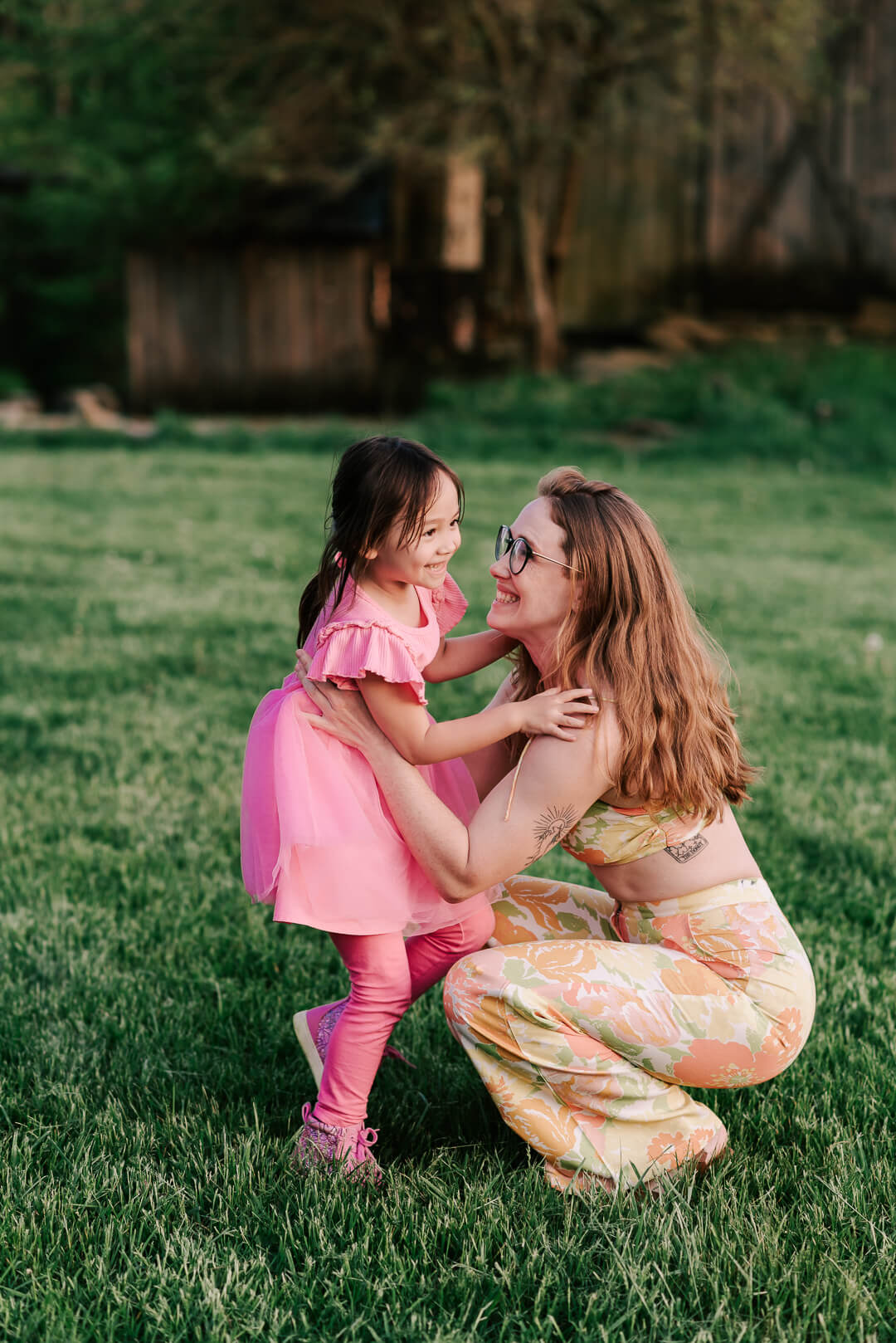 A happy mom in a floral print outfit plays with her toddler daughter in a pink dress in a lawn at sunset during some northern virginia kids activities