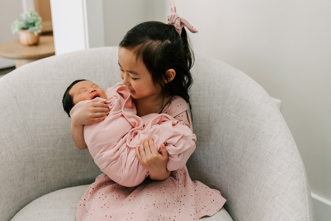 A toddler girl sits in a grey chair in a pink dress holding her sleeping newborn baby sibling