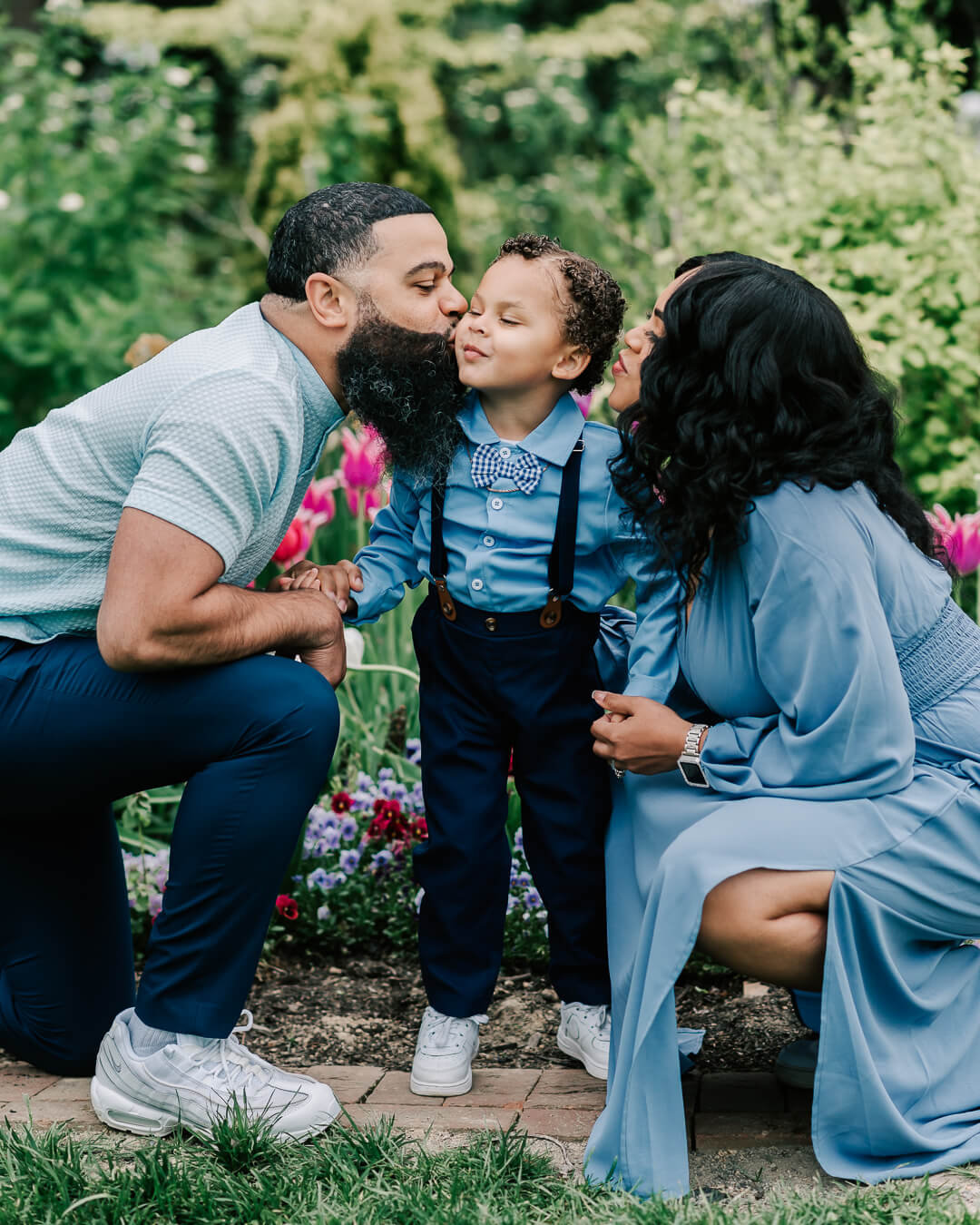 Happy parents kneel in a garden path while kissing their toddler son in a blue bowtie and suspenders