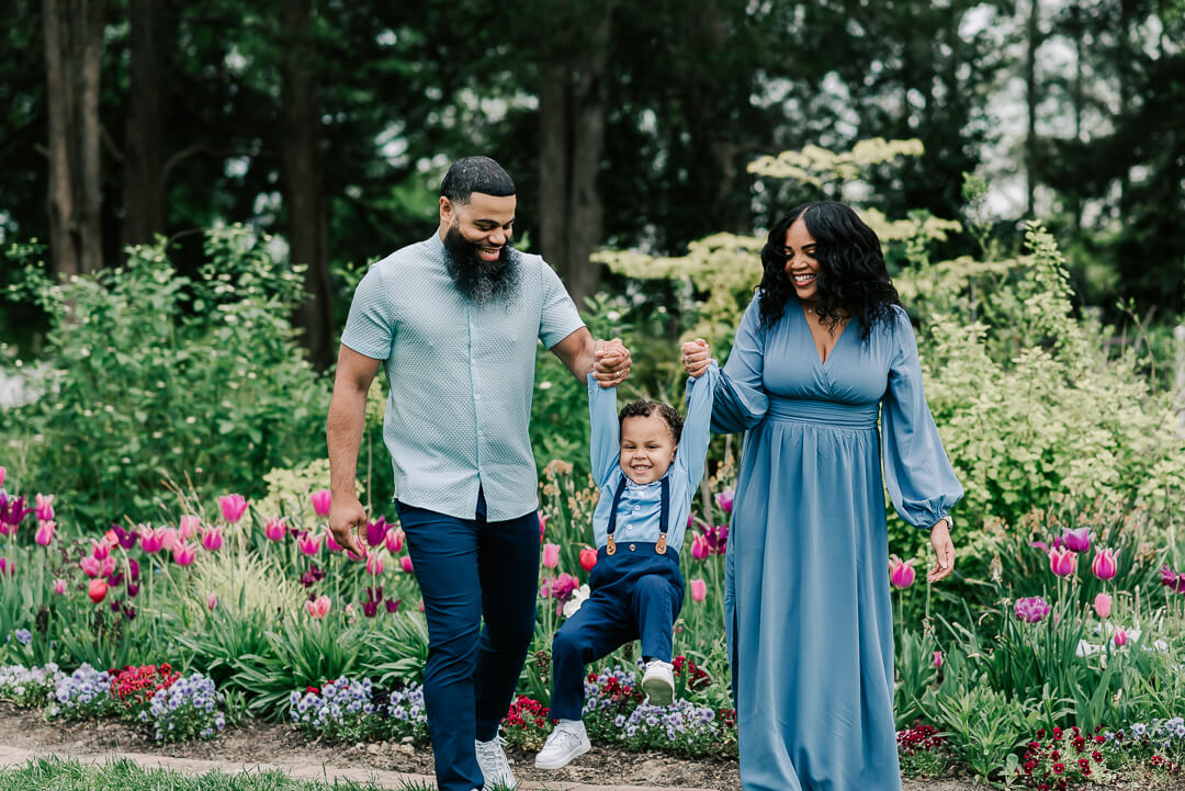 Mom and dad in blue swing their toddler son between them while walking through a flower garden before visiting splash pad fairfax va