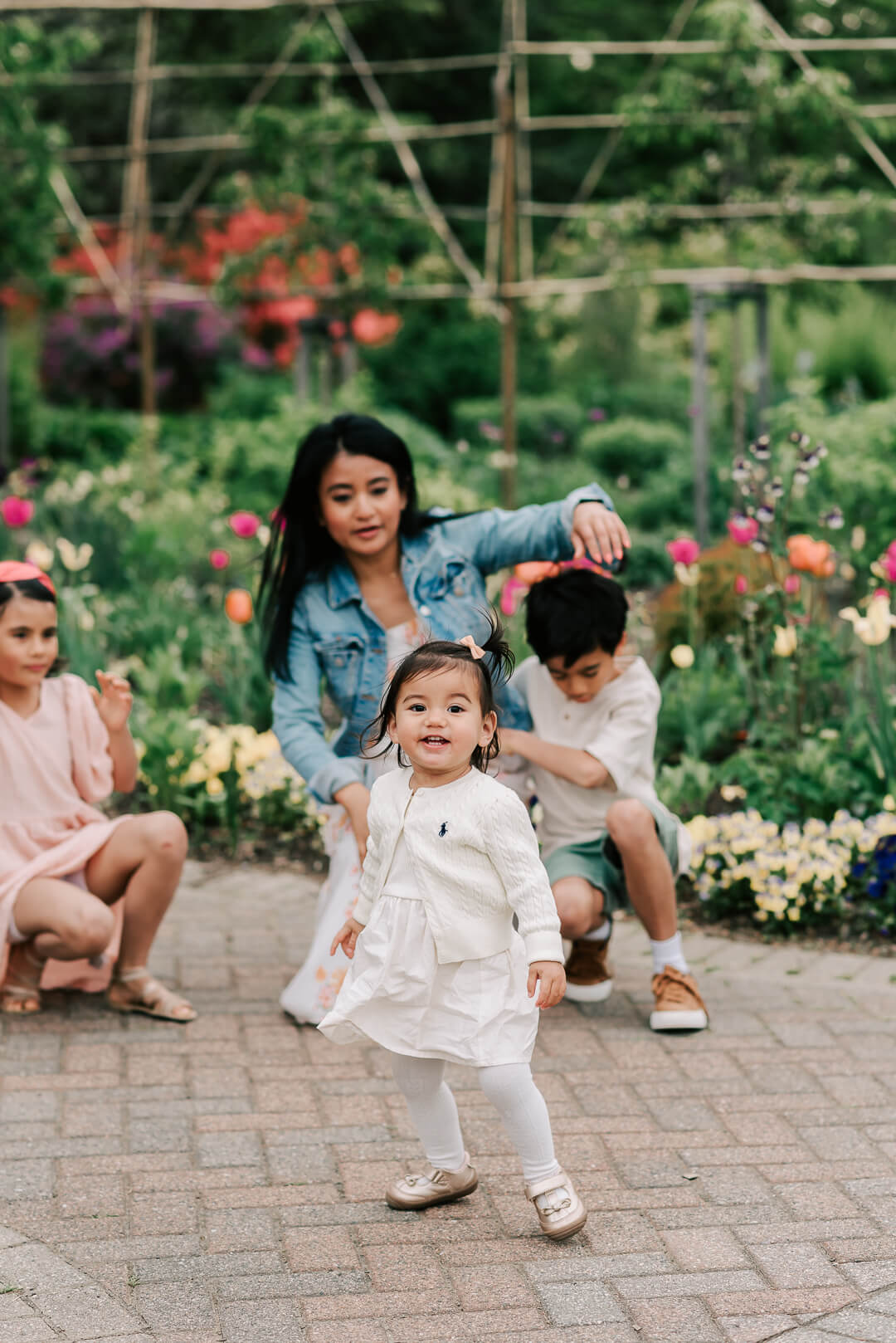 A mother kneels in a garden sidewalk while playing with her three toddler children before visiting trampoline park northern virginia