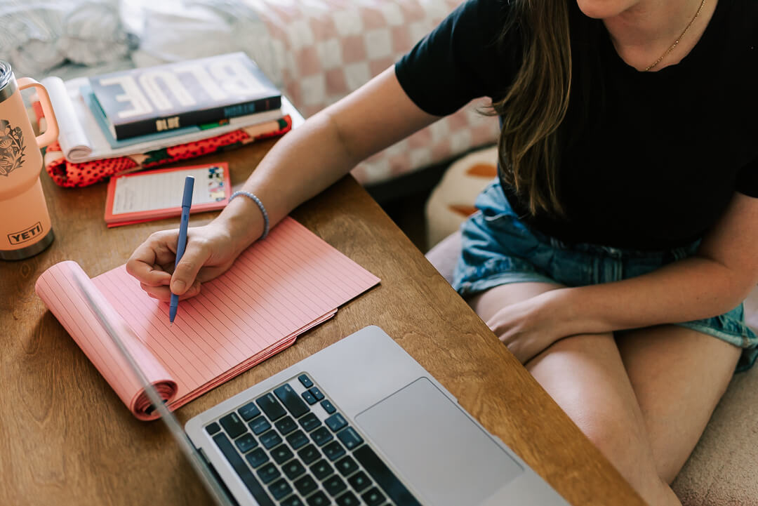 a woman writing notes at her desk with her laptop open